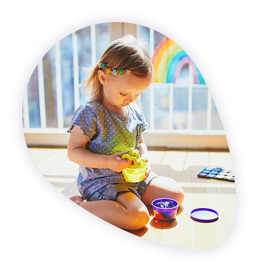 Kind mit vielen Haarklammern spielt auf dem Boden eines warmen Zuhauses, im Hintergrund ein Regenbogen aus Fingerfarben an einer Glasscheibe, symbolisiert die SWBB Fernwärmeversorgung für effiziente Energieversorgung.