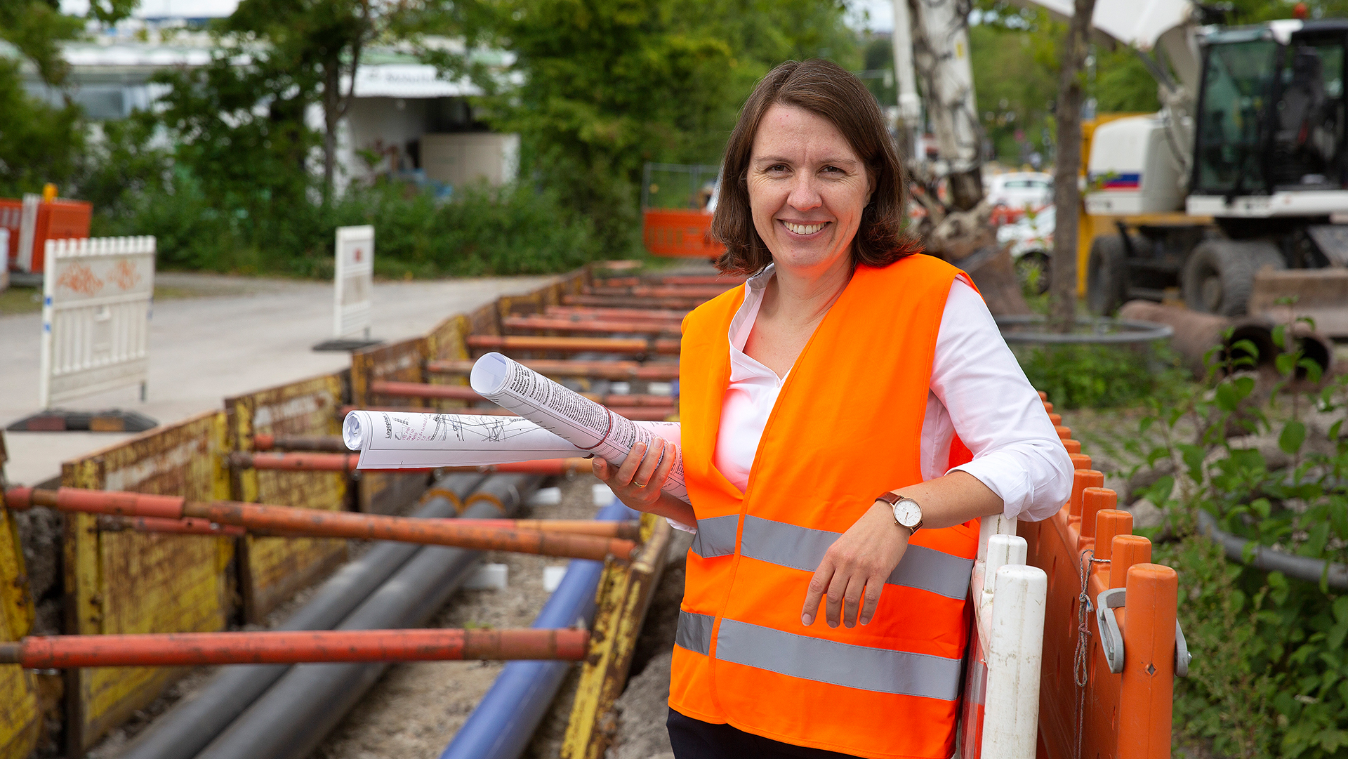 Bild einer lächelnden Frau mit Warnweste und Bauplänen in der Hand. Sie steht an Baustellenzäune gelehnt und im Hintergrund ist eine abgesperrte Baustelle mit offenen Rohren im freigelegten Boden zu sehen.