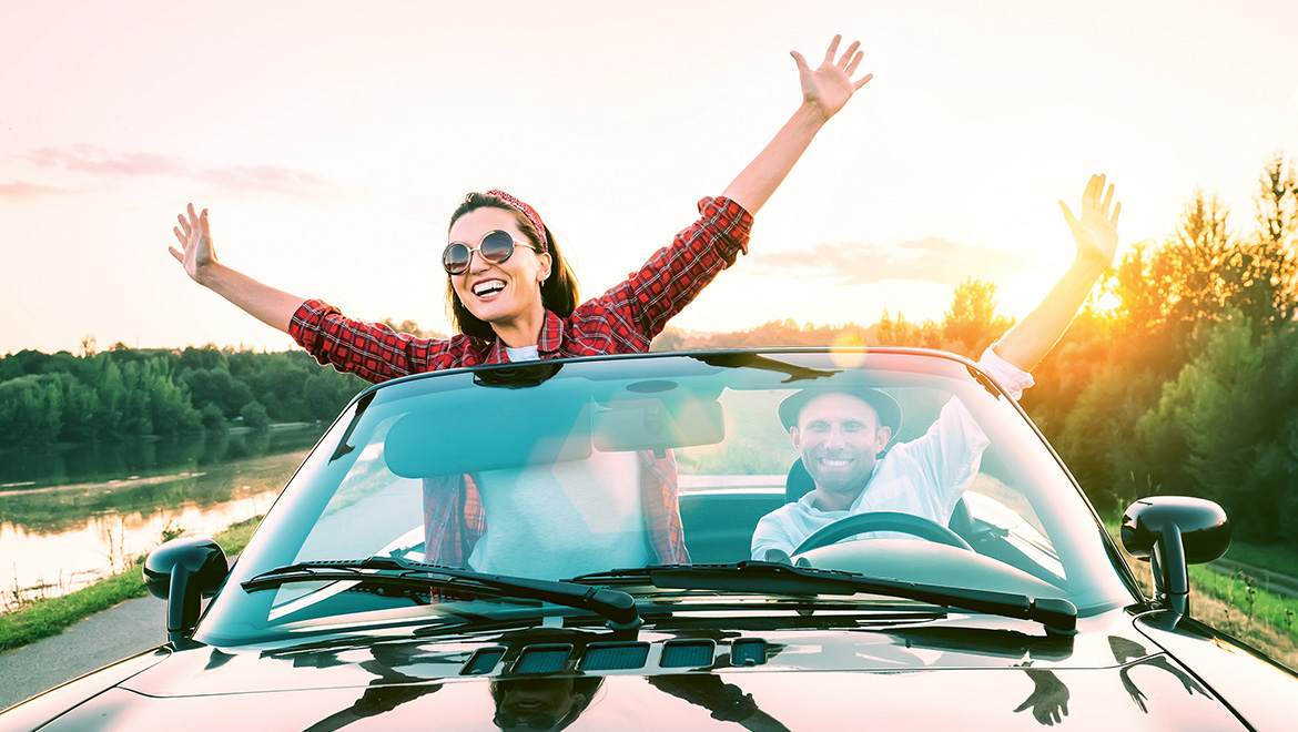 E-Mobil als Zweitauto in der Stadt: Bild von zwei glücklichen Personen in einem Cabrio. Im Hintergrund sind Bäume und eine Wasserfläche zu sehen.