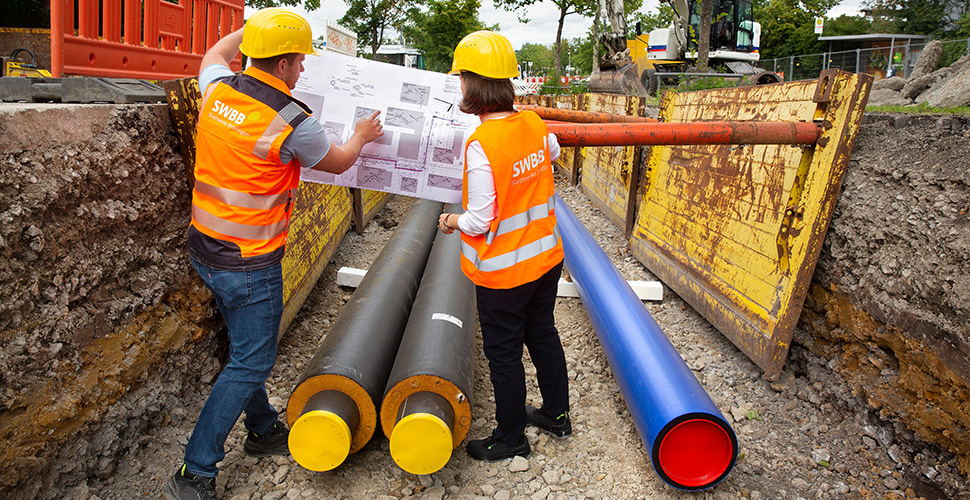 Verlegung von Fernwärmeleitungen in Böblingen, ein Mann und eine Frau mit Warnwesten stehen in einem Loch im Boden, in dem mehrere neue Rohre liegen. Sie betrachten einen Bauplan.