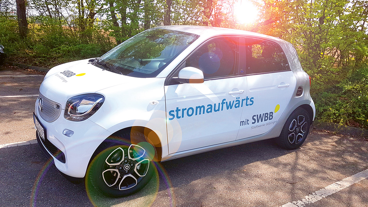 Weißer Elektrosmart der SWBB mit der Beschriftung "stromaufwärts" auf einem von Bäumen umgebenen Parkplatz.